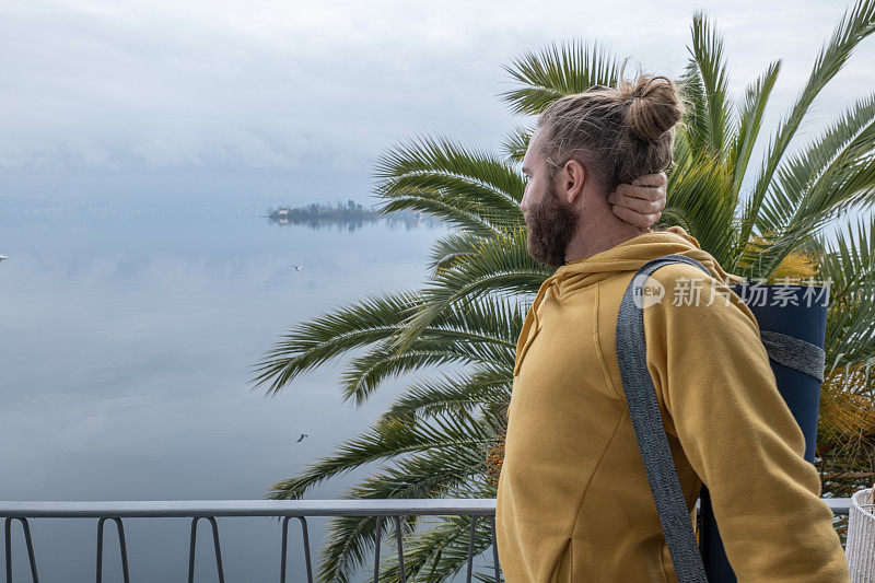 一个年轻人拿着瑜伽垫在她的阳台上沉思着湖和山的景色