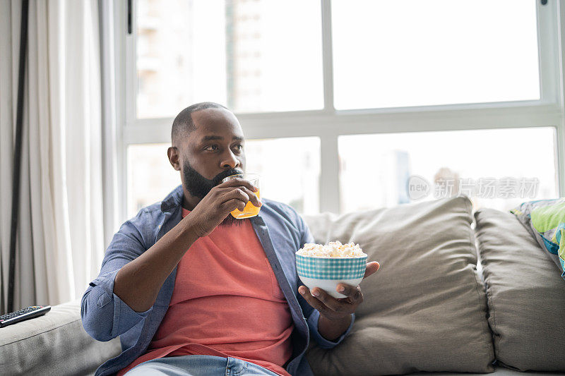 男人一边在家里看电视一边吃着爆米花，喝着果汁