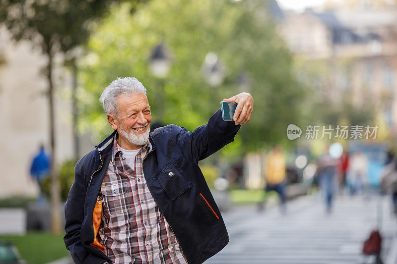 一个上了年纪的男人走在城市的街道上，用他的智能手机自拍。