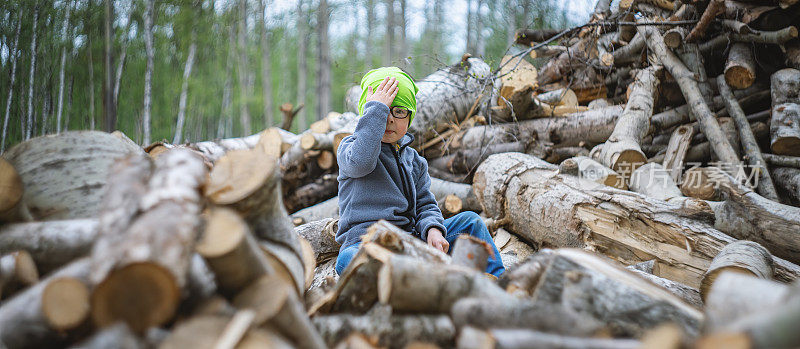 一个悲伤的小男孩坐在一堆被砍倒的树干上，原址是一片森林。Facepalm指的手势。城市扩张和城市化的生态问题。宽的形象。