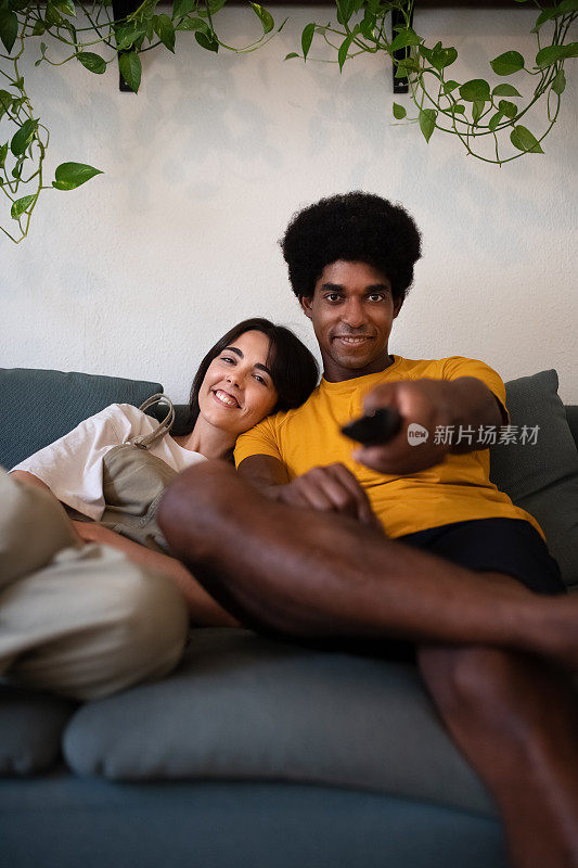 年轻的跨种族夫妇在家里看电视。非裔美国人用遥控器换台。垂直的图像
