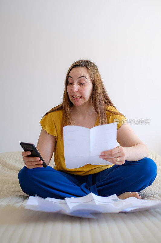 女人检查她的账单她的账单在她的床上，用手机计算器做数学，变得担心