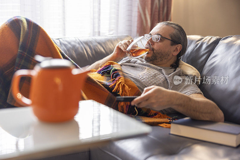 感觉不好。一个成熟的男人生病了，在温暖的格子里躺在沙发上休息。他正在用遥控器换电视频道，喝着热茶。