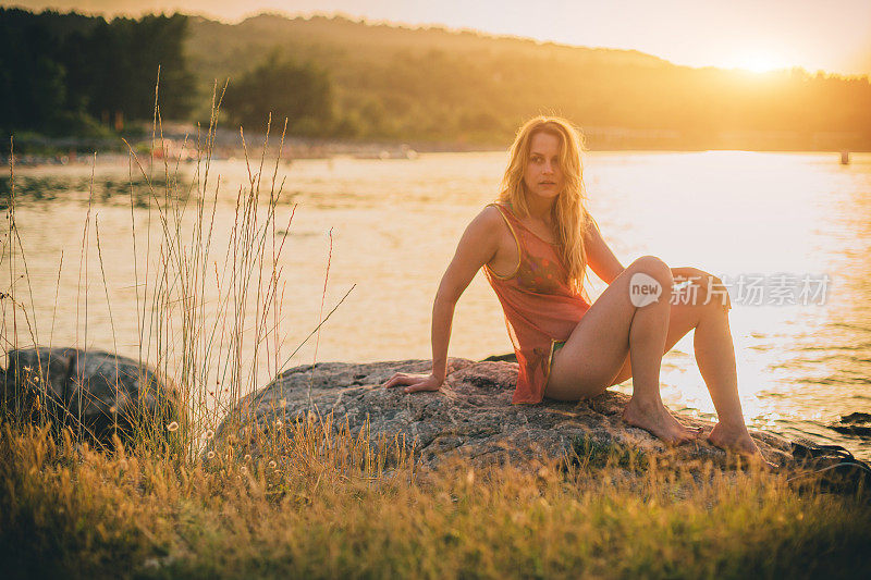 年轻女子坐在岩石上，看着美丽的日落和大海