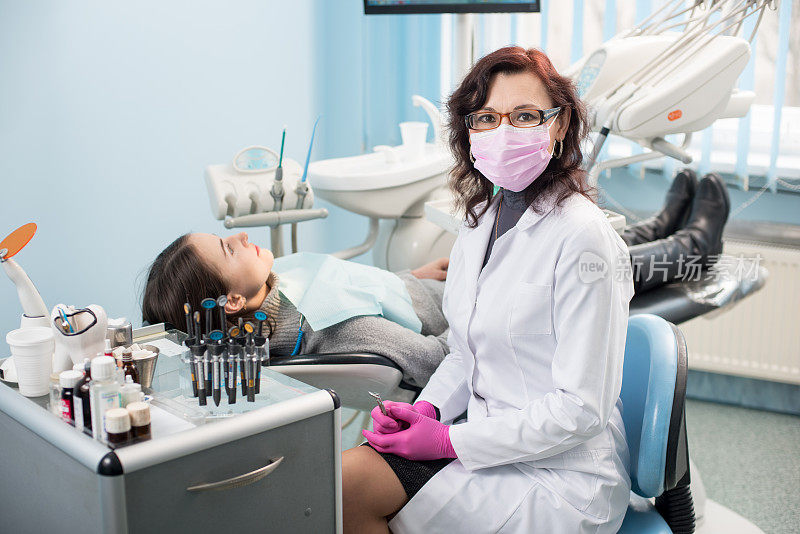 女牙医和女病人坐在牙科诊所的椅子上。医生戴眼镜，口罩，白色制服和粉红色手套。牙医。牙科设备
