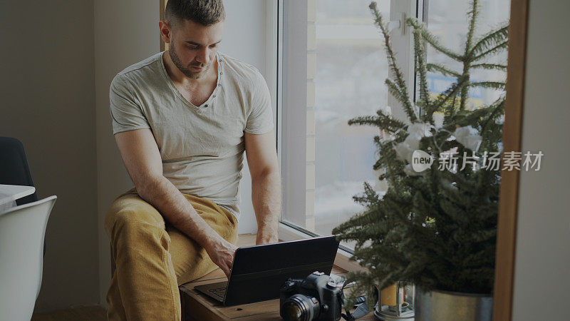 专注的男性摄影师拿着相机和笔记本电脑坐在家里的窗台上，把她的照片分享到社交媒体上