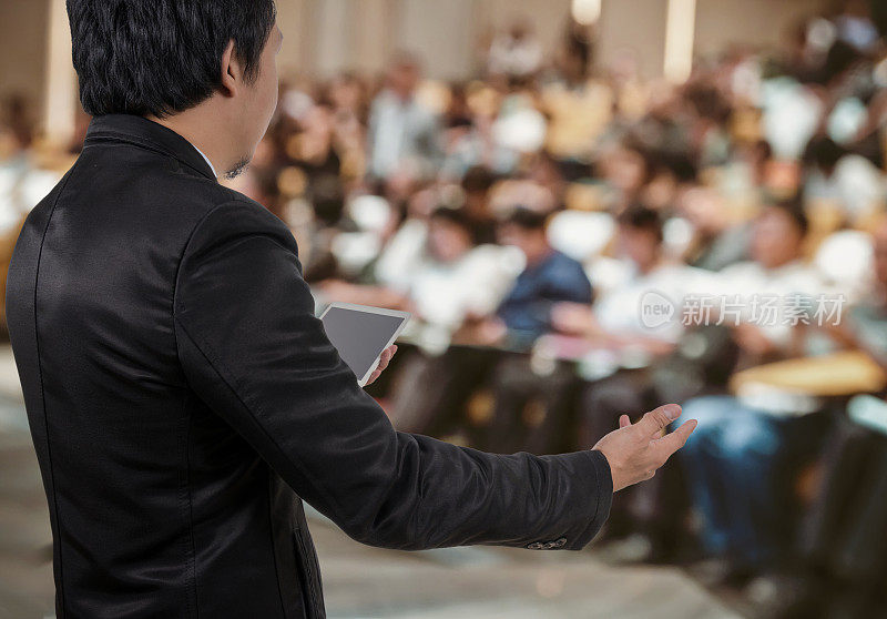 商务人士会议讲话者后视图上方是带有与会者背景、商务研讨会和教育理念的会议厅或会议室的抽象模糊照片