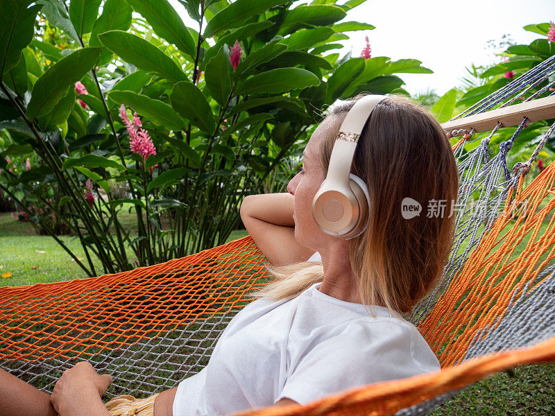 女孩听音乐在吊床-年轻女子戴着无线耳机和放松度假在热带花园