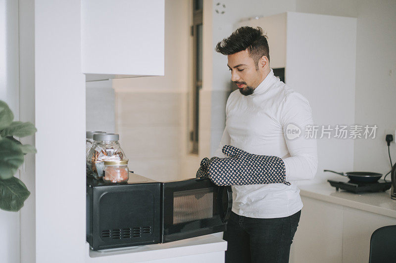 一名留着胡子的中东男子穿着高领衬衫，在厨房里戴着防护手套从微波炉里取出食物