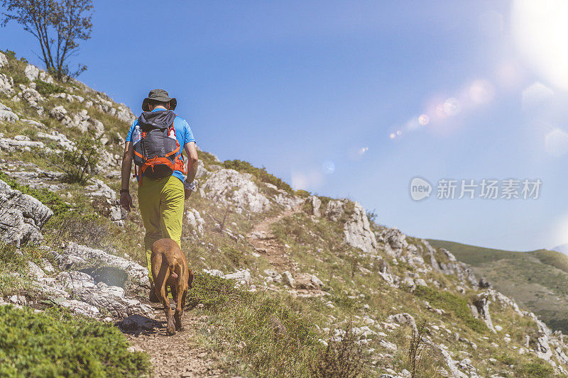 年轻的男性徒步旅行者走在岩石小径和他的棕色匈牙利维兹拉狗