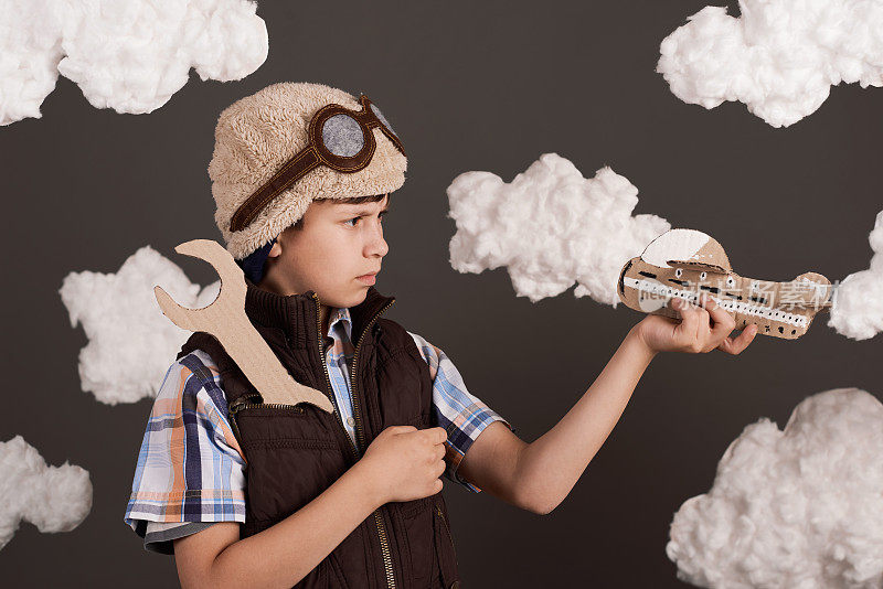 一个男孩在玩纸板飞机和扳手，梦想成为一名飞行员，穿着复古风格的夹克，戴着头盔和眼镜，棉絮云，灰色的背景，色调为棕色