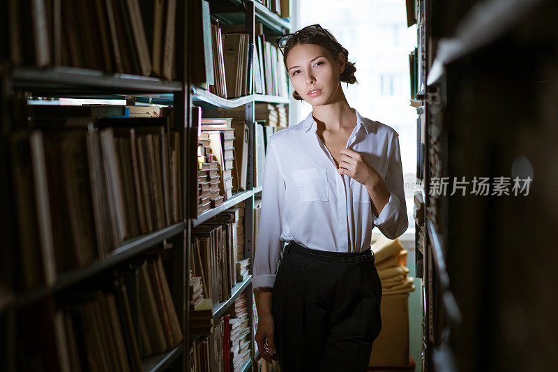 穿着白衬衫的漂亮女学生站在图书馆的两排中间，书架上放满了书。黑暗的照片