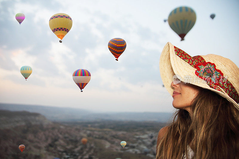 土耳其卡帕多西亚上空，一名戴着帽子的年轻女子在看热气球。令人印象深刻的景象