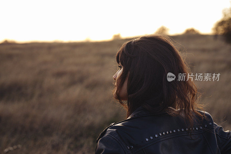 美丽的年轻女子穿着皮夹克享受日落在一个高草地