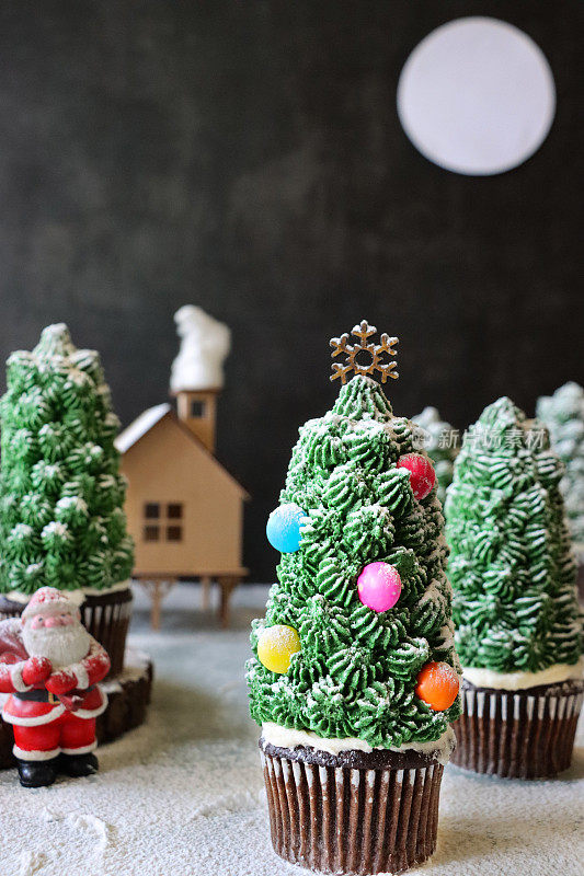 特写图像批量自制，圣诞树设计的巧克力纸杯蛋糕在纸杯蛋糕盒显示在雪，夜晚针叶林的场景，与圣诞老人和满月的模型房子