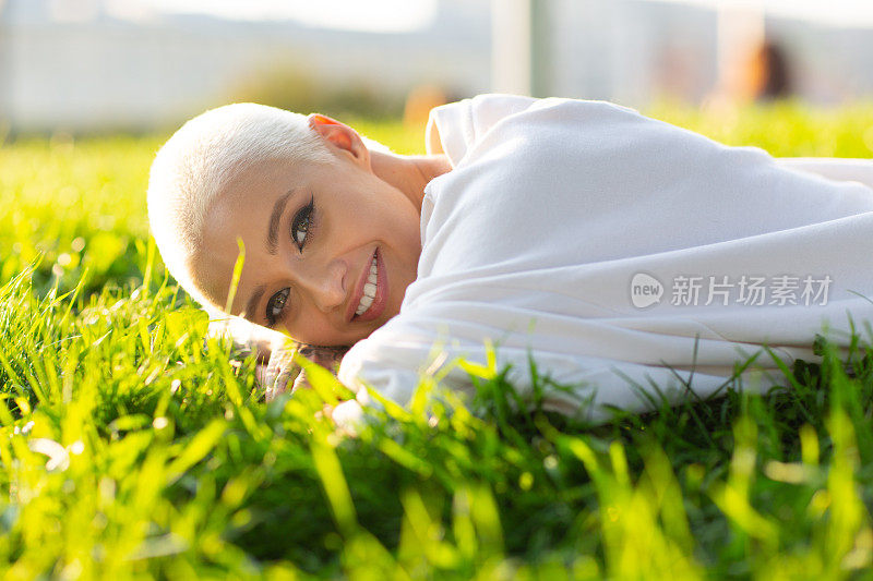 金发短发的千禧年轻女子在户外微笑着躺在草地上的画像。