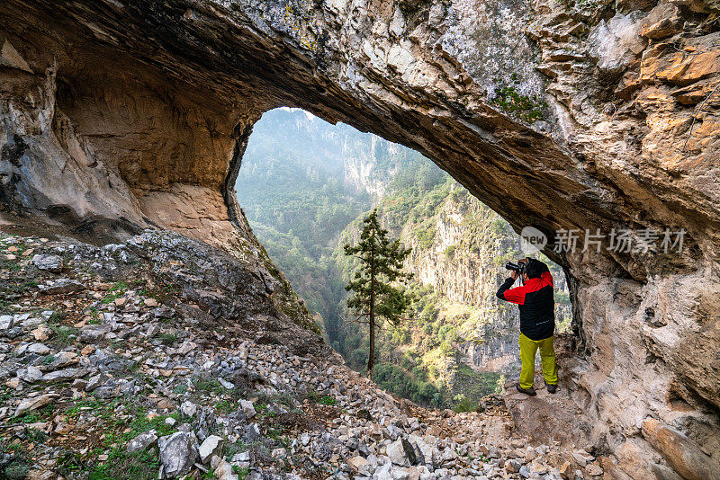 徒步旅行者摄影师在峡谷岩石拱门下，看着森林中的瀑布美景