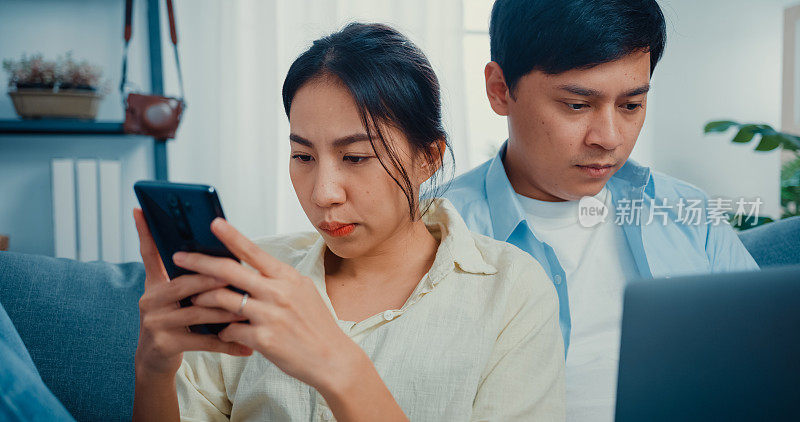 年轻的夫妇与休闲坐在沙发上妻子使用智能手机网上购物和丈夫使用笔记本电脑平板电脑在家里的客厅在家工作。