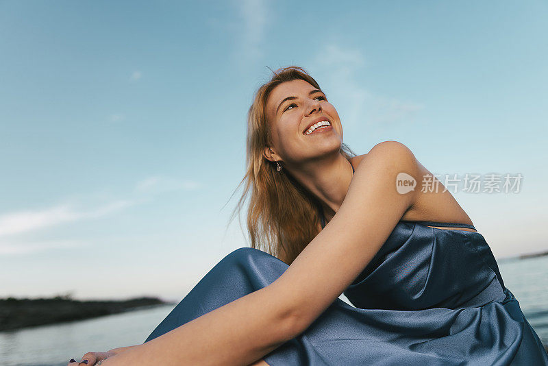 年轻的金发女子穿着蓝色的裙子，在夕阳下坐在海边。