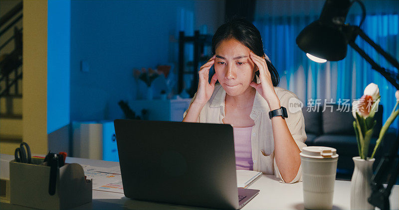 年轻的亚洲女性穿着正装衬衫坐在桌子前与笔记本电脑的屏幕上，认为感到压力，没有想法，使紧急报告会议，加班到午夜在家。