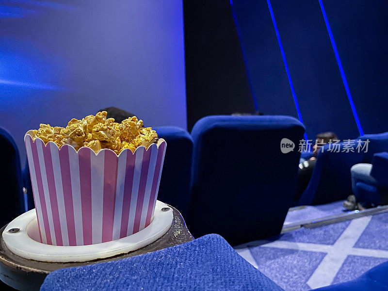 近距离图像的纸箱爆米花在电影院电影院座位杯托，电影零食食品浴缸，蓝色天鹅绒座椅，重点在前景