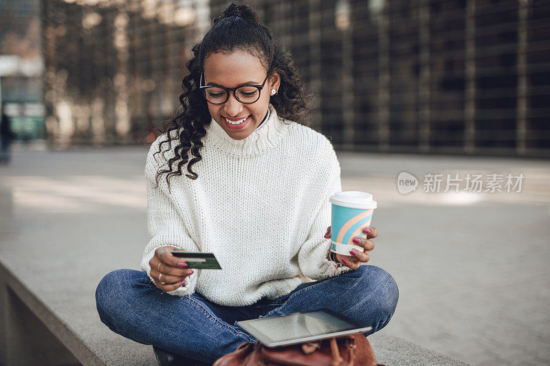 一位年轻的黑人妇女坐在木板路上的长凳上，用平板电脑在网上购物