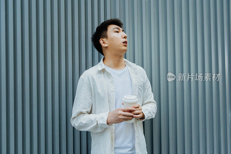 年轻的亚洲男性商人正在喝着咖啡，走在灰色的户外墙壁前——下班后喝一杯咖啡，促进健康，工作生活平衡