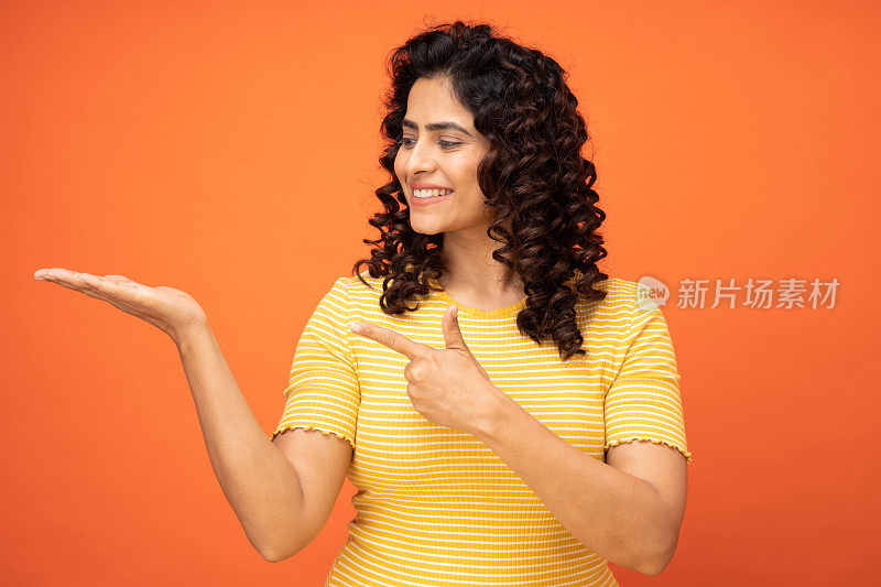 女孩漂亮的食指肖像，指示买家新产品在橙色背景的股票照片上张开的手