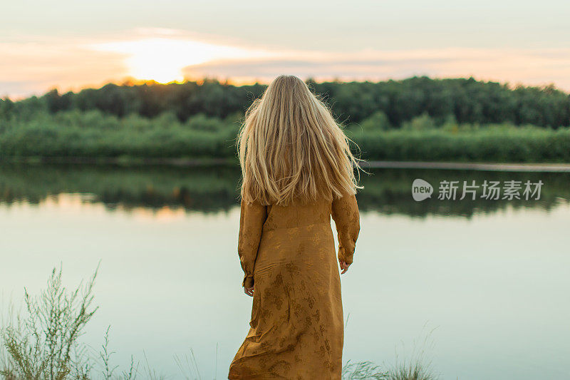 后面的观点。穿黄色衣服的金发女人躺在河岸上。夏天的一天。长头发的。沙子。