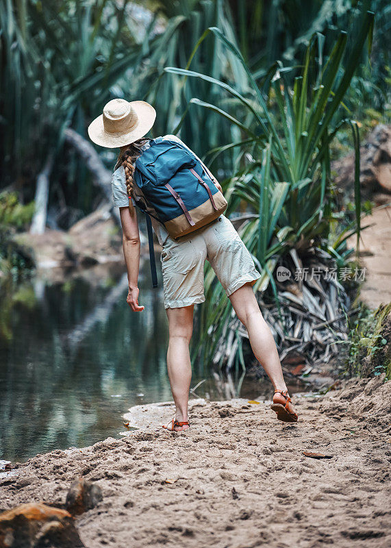 身穿米色衬衫、短裤、头戴草帽的年轻女子，肩上背着背包，在雨林丛林中徒步旅行，附近有一条小溪，从背后看过去