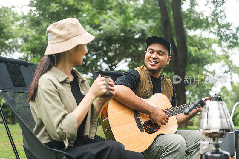 年轻的亚洲夫妇，男人和女人野餐坐在露营地户外花园。两个人在森林里露营，弹吉他，喝咖啡。旅游，放松，露营，度假，假日，周末的主题