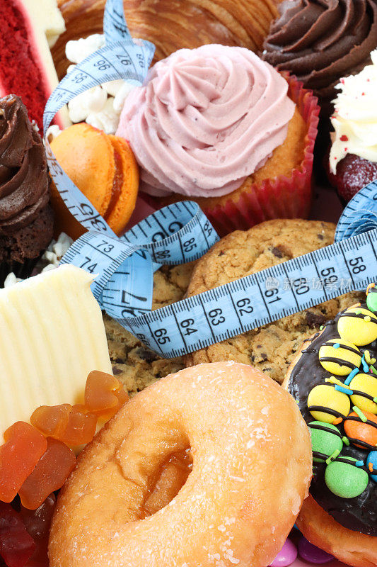 全画幅的蓝色卷尺上的甜垃圾食品，釉面环甜甜圈，软糖，红色天鹅绒蛋糕片，巧克力片饼干，蛋糕与黄油糖衣，马卡龙，牛角面包，重点前景，不健康的饮食观念