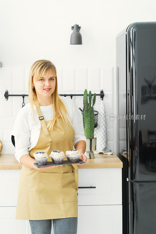 厨房里，冰箱旁边，一个金发的糖果师手里拿着巧克力纸杯蛋糕，上面有白奶油和浆果。特写镜头
