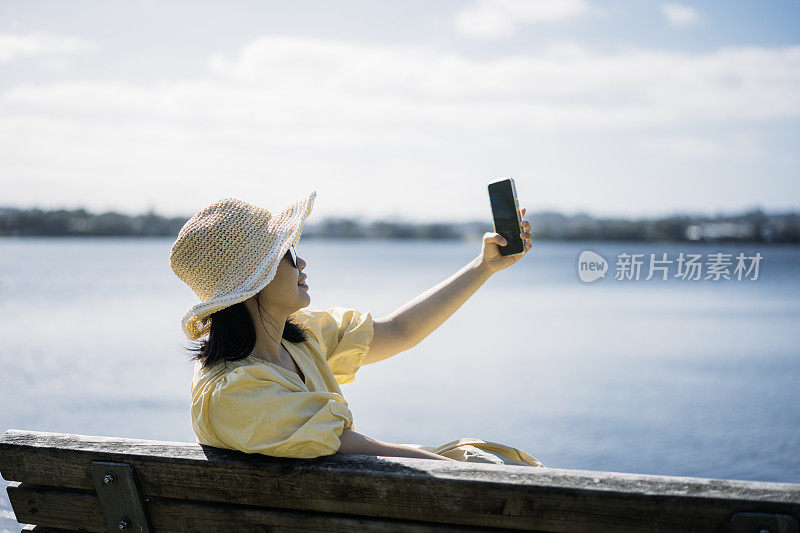 年轻的亚洲女子坐在湖边的长凳上，用智能手机自拍