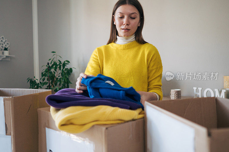 穿着黄色毛衣的年轻女子拿着衣服，在她的房间里放着捐款箱，表达着捐赠的概念