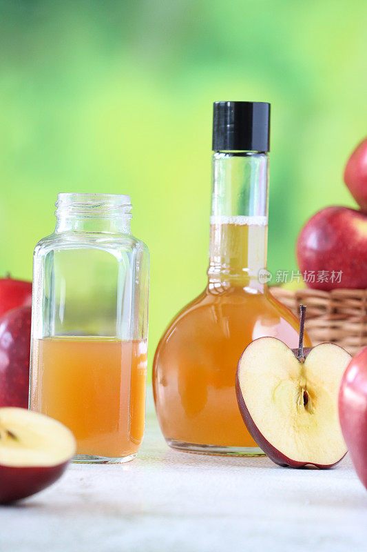 两个塑料瓶的特写图像，一个打开，一个关闭，装有苹果醋，柳条筐装有一堆红苹果，斑驳的绿色背景，重点前景，健康的生活方式理念