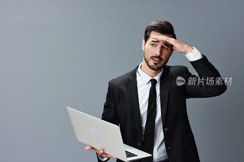 男人有胡子，黑发，商务头痛看着笔记本电脑屏幕上的手在灰色背景的西装。开展网上业务