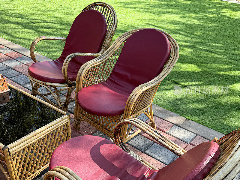 玻璃咖啡桌的特写图片，周围环绕着红色的藤扶手椅，红色和黑色铺砌的花园露台上的合成皮革垫子，人造绿色草坪草坪，重点在前景
