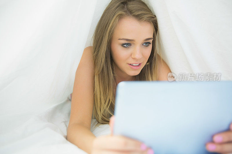 快活的金发女人在床单下使用平板电脑