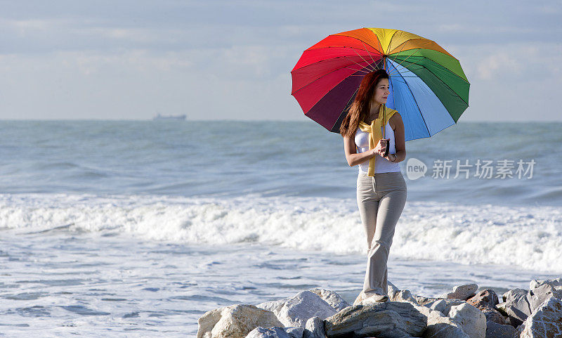 年轻女子拿着五颜六色的伞，在海边的岩石上