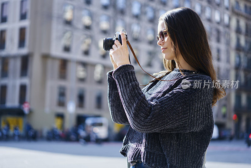女旅行者戴着太阳镜拿着老式相机从周末观光旅游拍照。专业摄影师在阳光明媚的日子拍摄城市背景建筑