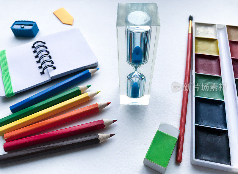 沙漏的特写和文具学校和创造力，绘画和工艺品(水彩颜料，彩色铅笔，橡皮，卷笔刀)与白色水彩纸上的文字空间