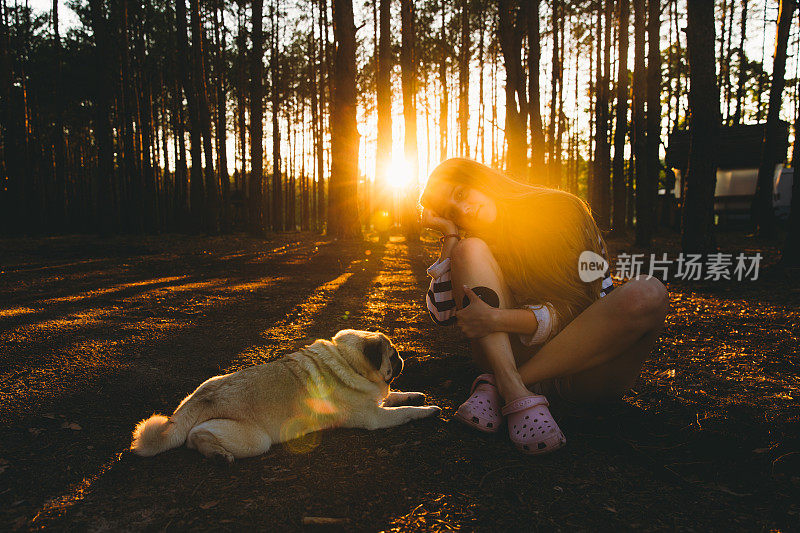一个年轻的女人和她的狗在松树林里遇见了明亮的日落