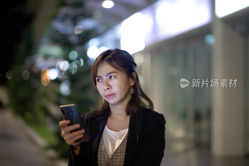 亚洲中国女人看着智能手机与担忧的表情脸在夜晚的街道上。