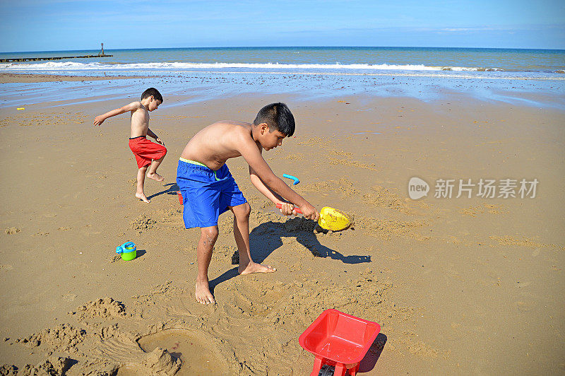 在一个阳光明媚的夏日，两个印度亚洲男孩用沙滩玩具铲沙子