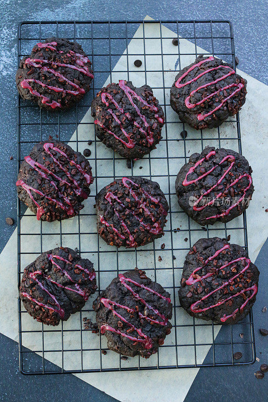 一排排新鲜烘焙的，自制的，巧克力屑黑森林饼干，装饰着粉红色的水糖霜，冷却在电线上，金属架上面的防油纸羊皮纸，聚焦前景