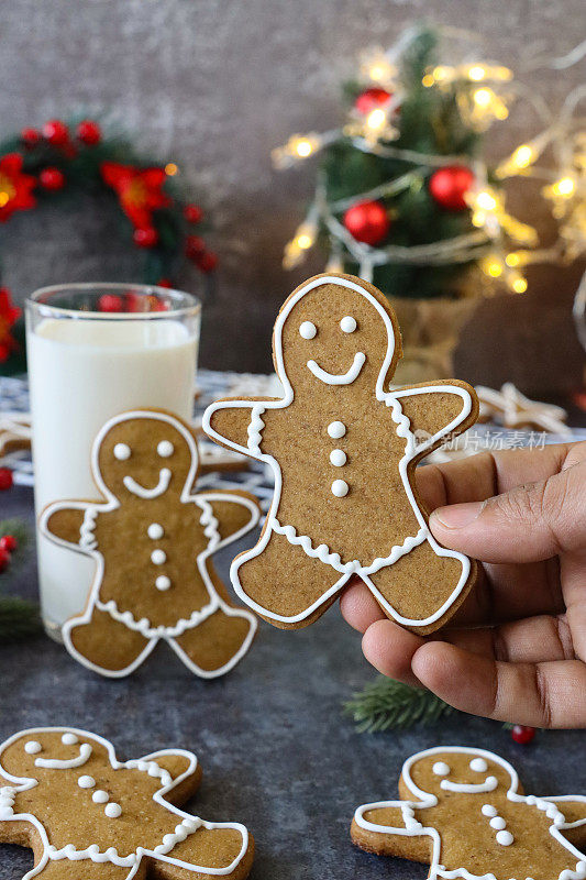 无法辨认的人拿着姜饼人饼干，用白色皇家糖霜装饰，喝牛奶，迷你圣诞树，红色和金色的装饰物，花环，灰色背景，家庭烘焙