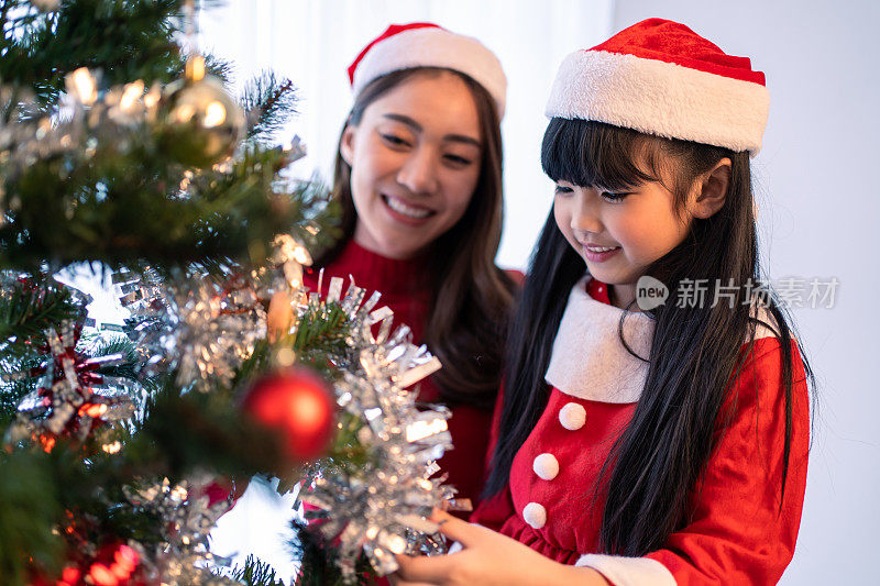 亚洲可爱的家庭，母亲和女儿装饰圣诞树。年幼的小女儿感到高兴和兴奋，与父母一起在家中庆祝节日圣诞节感恩节聚会