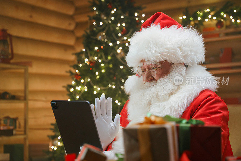 圣诞老人坐在装饰有圣诞树的小木屋里的一张桌子旁，用他的数码平板电脑向某人进行视频问候，前景是礼物