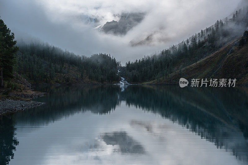 山间小溪从森林丘陵流入冰湖。雪山岩在雾中清场。小河和针叶树倒映在平静的高山湖上。清晨宁静的雾蒙蒙的风景。